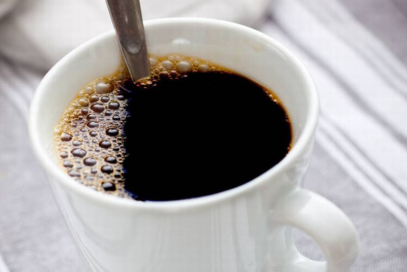 Raidallisen pöytäliinan päällä valkoinen kuppi, jossa on kahvia ja lusikka. 