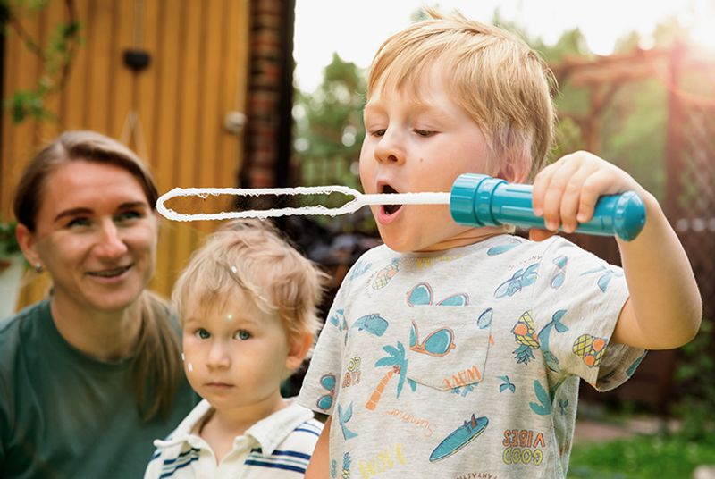Kuvassa on kaksi alle kouluikäistä poikaa, ja heidän isänsä. Toinen poika puhaltaa isoja saippuakuplia.