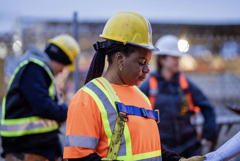 Naispuolinen rakennustyöntekijä työmaalla, taustalla toisia työntekijöitä.