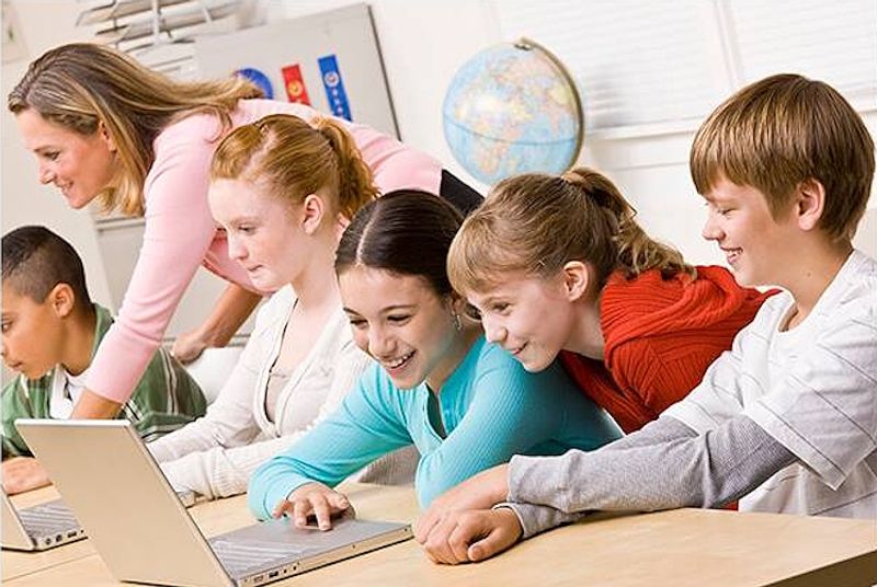 Iloisia koululaisia luokassa pöydän ääressä, osalla edessään kannettava tietokone.