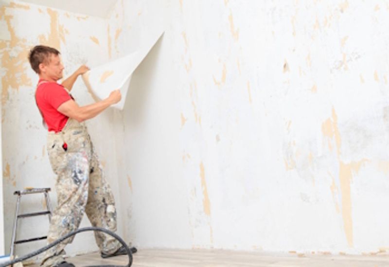 Valokuvassa mies työhaalareissa repimässä vanhaa tapettia seinästä.