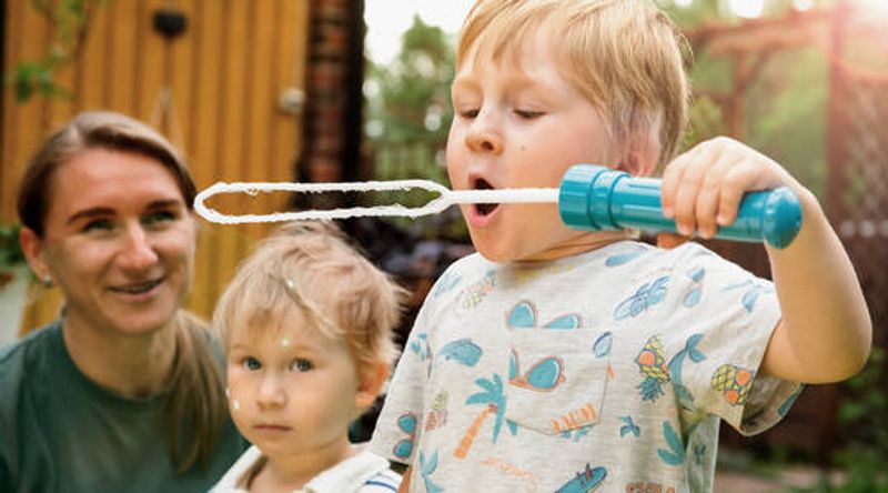 Kuvassa äiti ja kaksi poikaa. toinen poika puhaltaa saippukuplia isolla saippuakuplavälineellä.