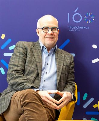 TIlastokeskuksen pääjohtaja Markus Sovala istuu hymyillen keltaisella tuolilla. Sovalan takana on Tilastokeskuksen 160-vuotisjuhlan logo.