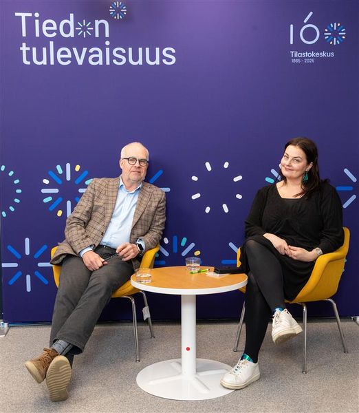 Tilastokeskuksen pääjohtaja Markus Sovala ja valtiosihteeri Kristiina Kokko istuvan sinisen hymyillen tuoleilla. Tuolin takana on sininen tausta, jossa lukee "Tiedon tulevaisuus" ja "160 Tilastokeskus 1865–2025".