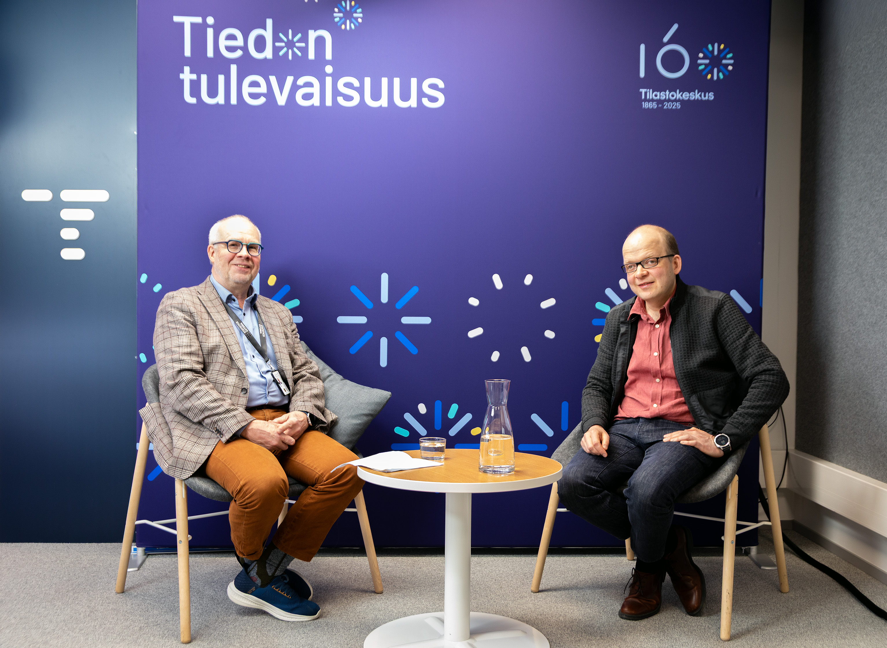 Tilastokeskuksen pääjohtaja Markus Sovala ja Helsingin Sanomien kehitysjohtaja Esa Mäkinen istuvat hymyillen Tilastokeskuksen juhlavuositaustan edessä. 